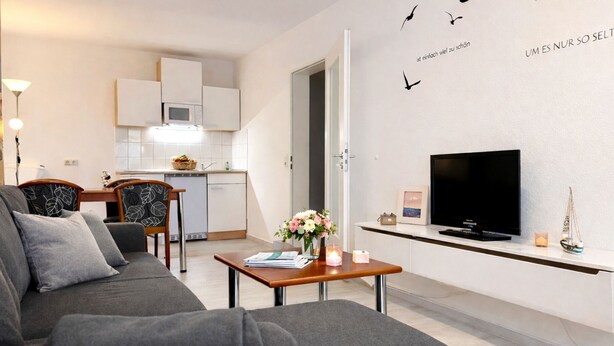 A modern living room with a grey sofa, a wooden coffee table, and a flat-screen TV. A kitchen with white cabinets and a microwave is visible in the background. - Welcome to “Donnerkeil” – your cozy seaside hideaway. (Ostseebad Binz)