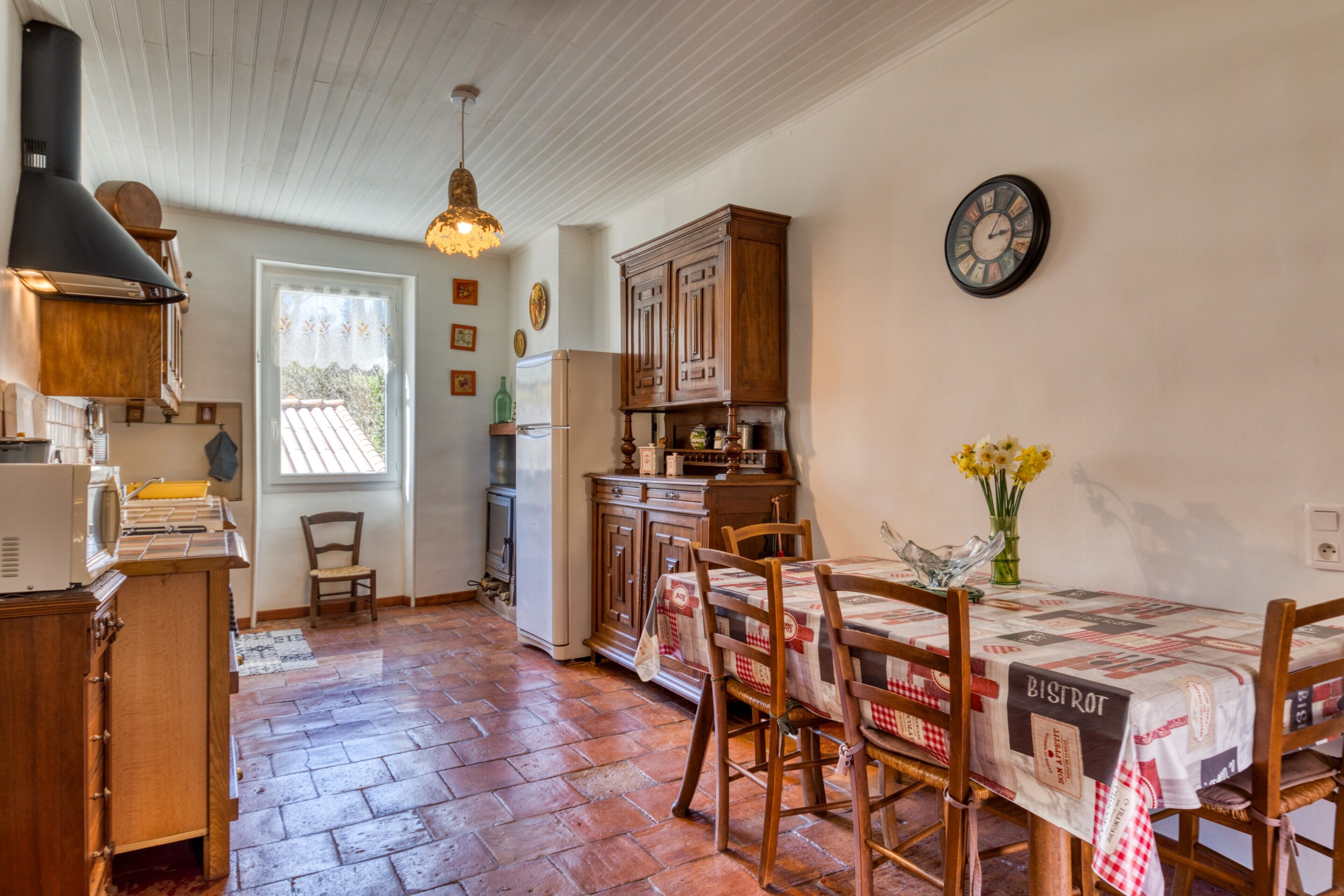 A kitchen with wooden cabinets, a dining table with chairs, a clock on the wall, and a window with a view.