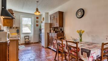 A kitchen with wooden cabinets, a dining table with chairs, a clock on the wall, and a window with a view.
