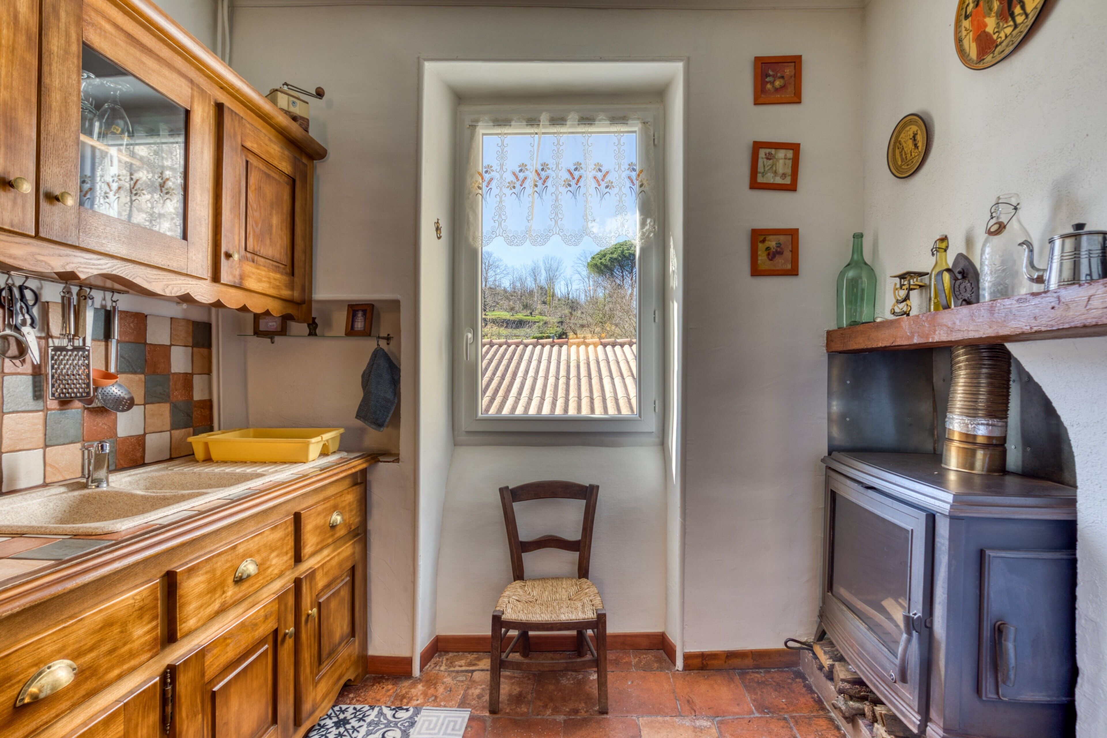 Une cuisine avec des meubles en bois, une fenêtre ornée de rideaux en dentelle, une chaise et un poêle.