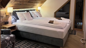 Un dormitorio con techo inclinado, una cama grande, una mesita de noche, una silla y una alfombra estampada.
