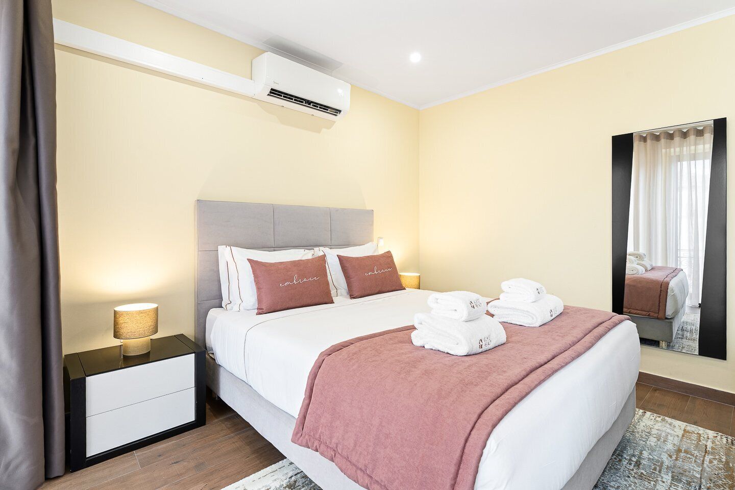Habitación de hotel con cama, mesitas de noche, aire acondicionado, espejo y ventana con cortinas.
