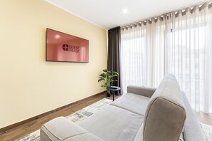 A modern living room with a sofa, a TV mounted on the wall, a potted plant, and a view through sheer curtains. - GuestReady - Poveiros Flats (Porto)
