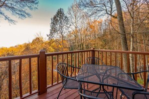 Een houten terras met een metalen tafel en stoelen, met uitzicht op een bebost gebied met herfstkleuren.