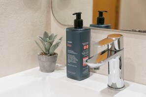 Ein Badezimmer mit einem weißen Waschbecken, einer grauen Flasche Anyah Körperlotion, einer kleinen Topfpflanze und einem chromfarbenen Wasserhahn.