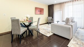 Een moderne woonkamer met een glazen eettafel, grijs gestoffeerde stoelen, een grijze bank en een groot raam met dunne gordijnen.