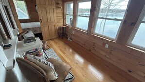 Interior de una cabaña con paredes y piso de madera, un escritorio con una silla, una ventana que ofrece vistas a un lago y una puerta que da a otra habitación.