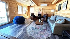 Interior acogedor de una cabaña con piso de madera, un tapete redondo, un sofá, una silla y un sillón de peluche.