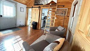 Una habitación amplia con piso de madera, un sofá con cojines estampados, una escalera y una cocina visible a través de una puerta corrediza.