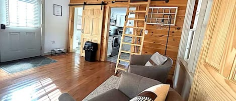 A spacious room with wooden flooring, a sofa with patterned cushions, a ladder, and a kitchen area visible through a sliding door.