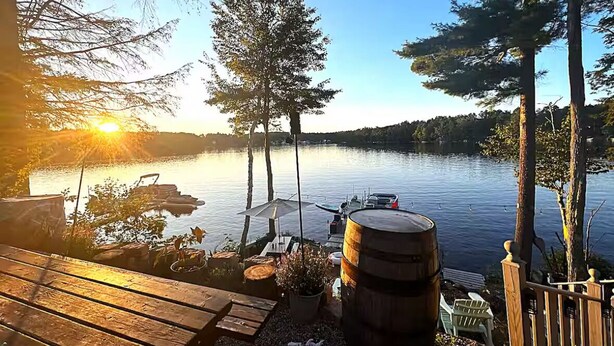 Terraza junto al lago con bancos de madera, un barril y vistas al lago y al atardecer.
