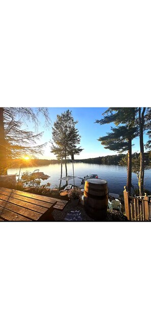 A lakeside deck with wooden benches, a barrel, and a view of the lake and sunset. - Maine Escape on Mousam Lake - Pet Friendly (Shapleigh)
