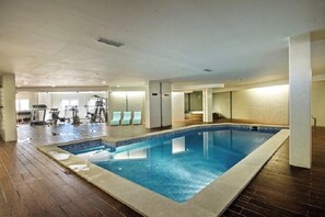 An indoor swimming pool with a wooden floor surrounding it, a fitness area with exercise equipment, and a seating area with chairs. - Excellent Private Penthouse with lots of space (Lagos)