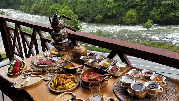 Een tafel met diverse gerechten en een theepot, voor een achtergrond van een bebost rivierlandschap.