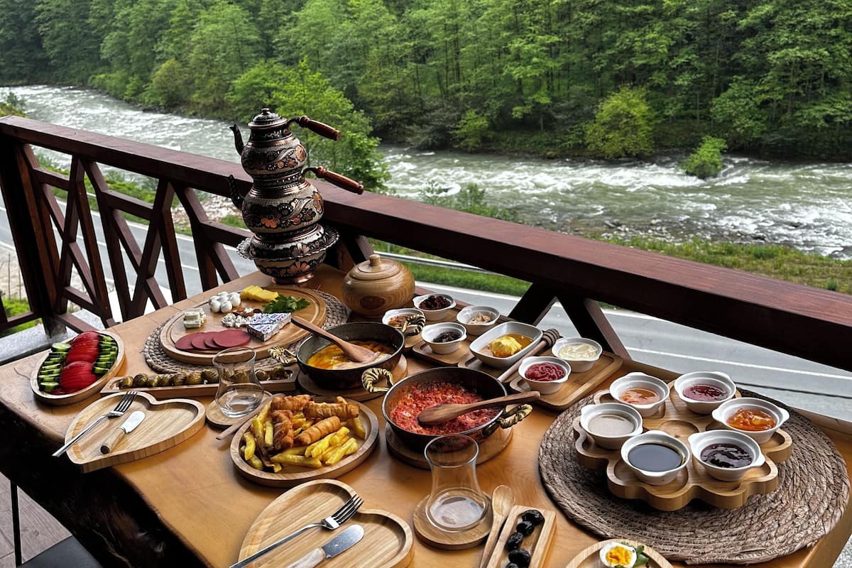 A table with a variety of foods and a teapot, set against a backdrop of a forested river.
