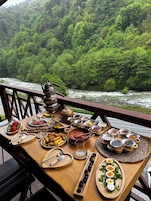 Una mesa con diversos alimentos y una tetera, frente a un río rodeado de bosque.