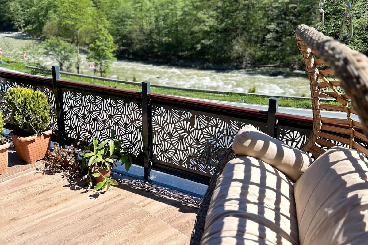 A wooden deck with a wicker sofa and potted plants, overlooking a river and forested area.