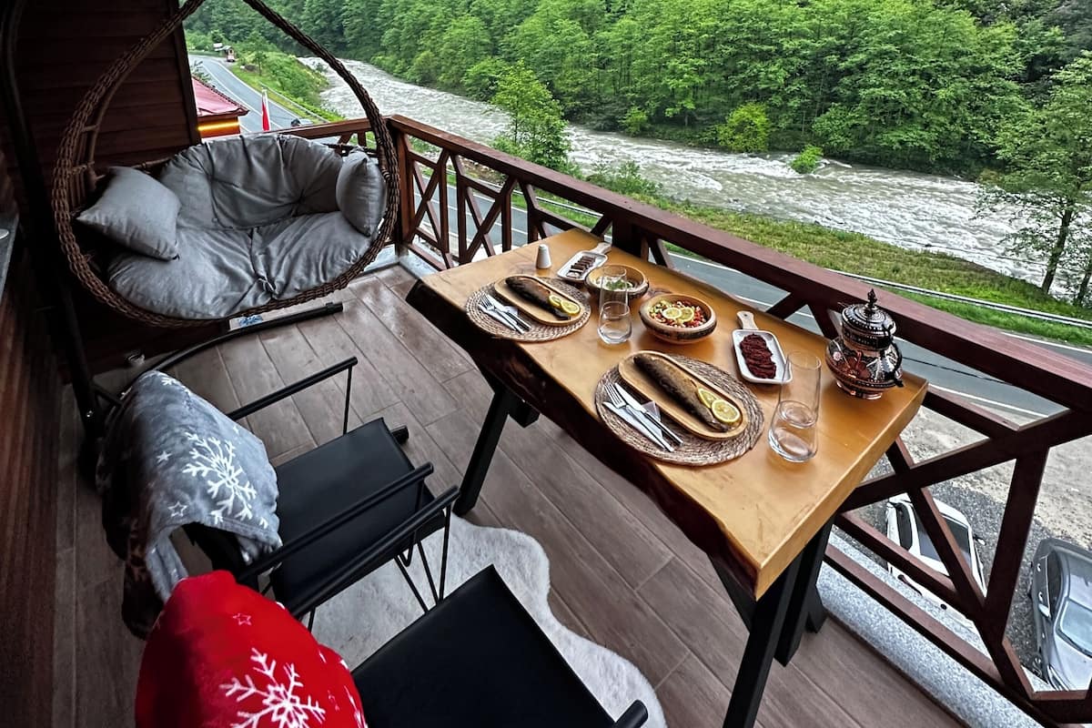 A dining area with a table set for two, a wicker chair, a cushioned bench, and a view of a river and forest.