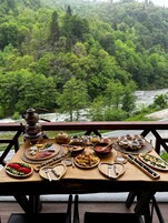 Una mesa dispuesta con diversos alimentos y un calentador de agua, con vistas a una zona boscosa con un río.