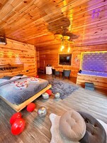Interior de una cabaña de madera con una cama, un televisor, una zona de comedor y una chimenea.
