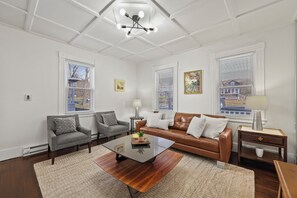 A living room with a brown leather sofa, two grey armchairs, a glass coffee table, and a wooden side table with a lamp.