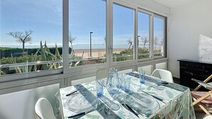 A dining area with a table set for four, large windows with a view of the beach, and a black cabinet to the right. - LA PLAGE Roses / Roses - INTERimmo (Rosas / Roses)