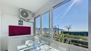 A dining area with a table set for two, a red partition, and a view of the sea and hills. - LA PLAGE Roses / Roses - INTERimmo (Rosas / Roses)