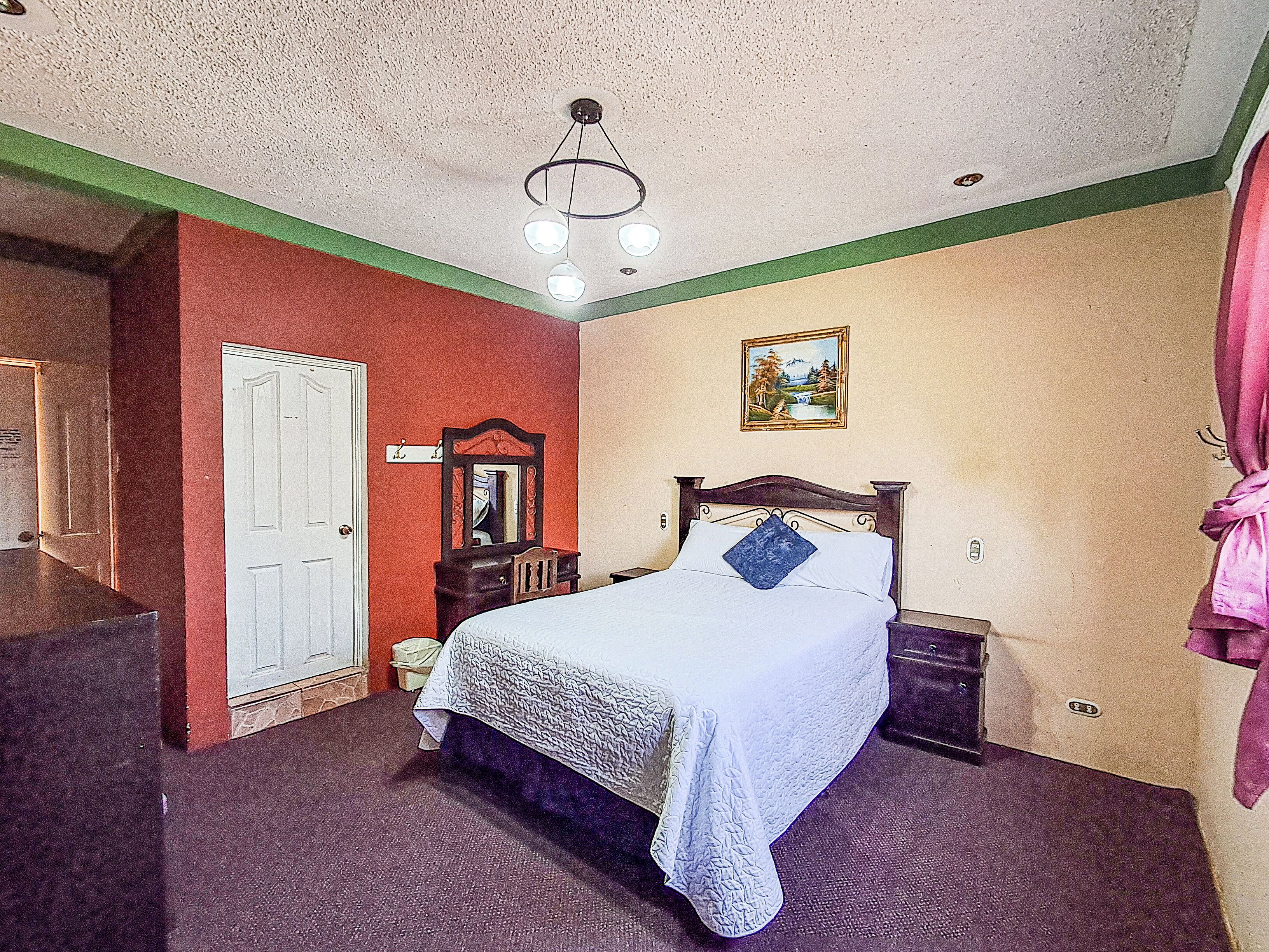 A bedroom with a bed, a dresser, a mirror, a painting, and a ceiling light fixture.