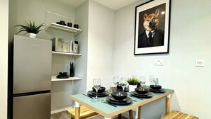 Uma área de refeições com uma mesa posta para quatro pessoas, uma geladeira, uma estante com utensílios de cozinha e um quadro na parede.