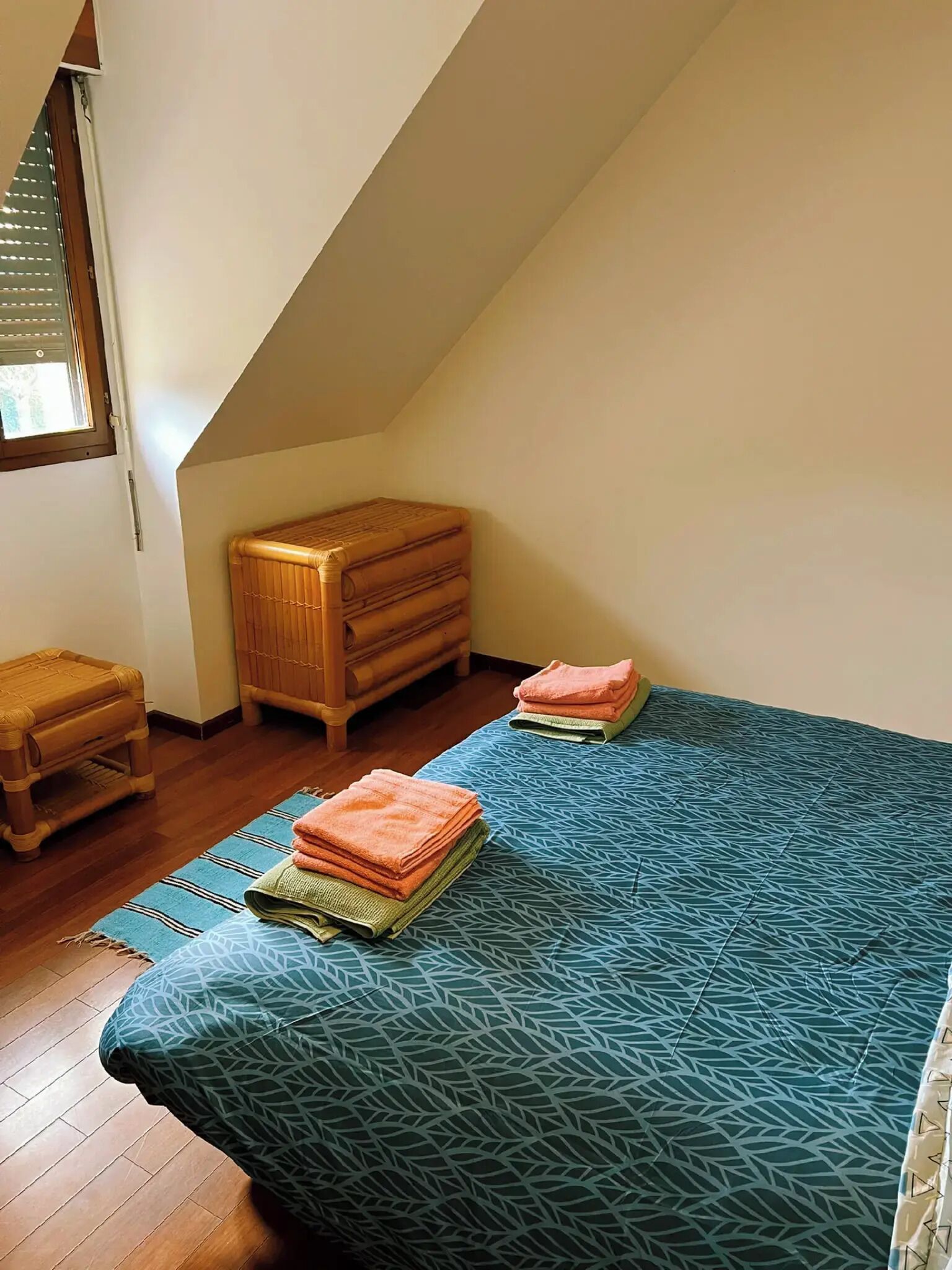 A small, cozy room with a sloped ceiling, a wooden chest of drawers, a small wooden stool, and a bed with a patterned blue bedspread.