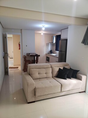 A living room with a beige sofa, a dining table, and a kitchen area with a refrigerator and stove. - Apartment on Palmas beach (Governador Celso Ramos)