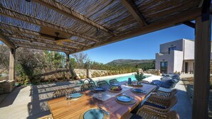 Un espace de restauration en plein air avec une table en bois et des chaises en osier, offrant une vue sur une piscine et un bâtiment moderne.