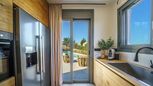 Une cuisine moderne dotée d’un réfrigérateur en acier inoxydable, de meubles en bois, d’une grande fenêtre et offrant une vue sur une piscine et un espace de détente extérieur.