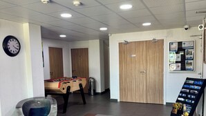Un hall d’entrée avec un baby-foot, un tapis bleu sur lequel est inscrit le texte « ADRECHAS MERCANTOUR », un espace de détente circulaire et une porte avec une pancarte.