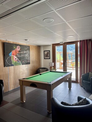 Ein Billardtisch in einem Raum mit einer Decke mit geometrischem Muster, einem Gemälde an der Wand und einem Fenster mit Blick auf eine Berglandschaft.