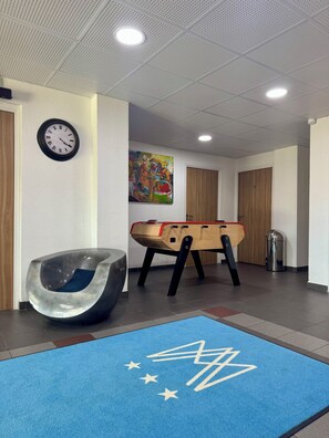 A room with a blue mat, a clock, a colorful painting, a wooden foosball table, and a modern chair. - ADRECHAS MERCANTOUR (Valdeblore)