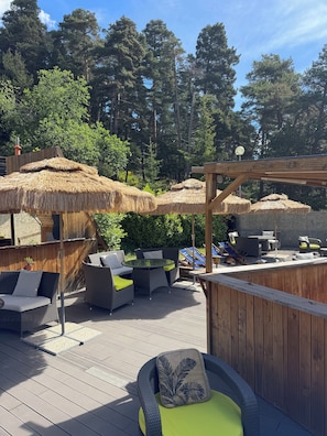 A wooden deck with outdoor furniture, including wicker chairs and a table, surrounded by thatched umbrellas and a wooden pergola. - ADRECHAS MERCANTOUR (Valdeblore)