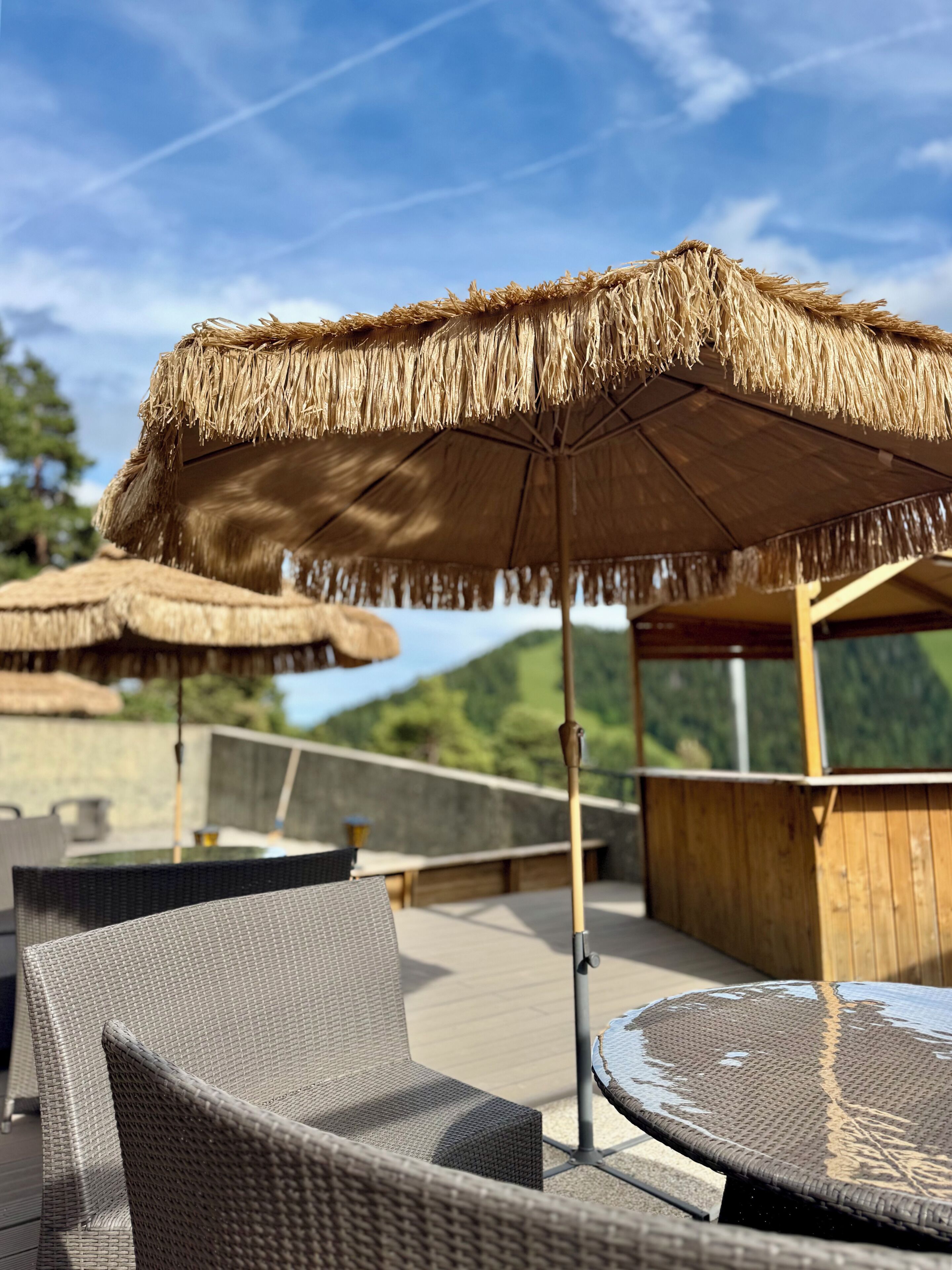 Outdoor seating with wicker chairs and thatched umbrellas, a wooden deck, and a mountainous backdrop.