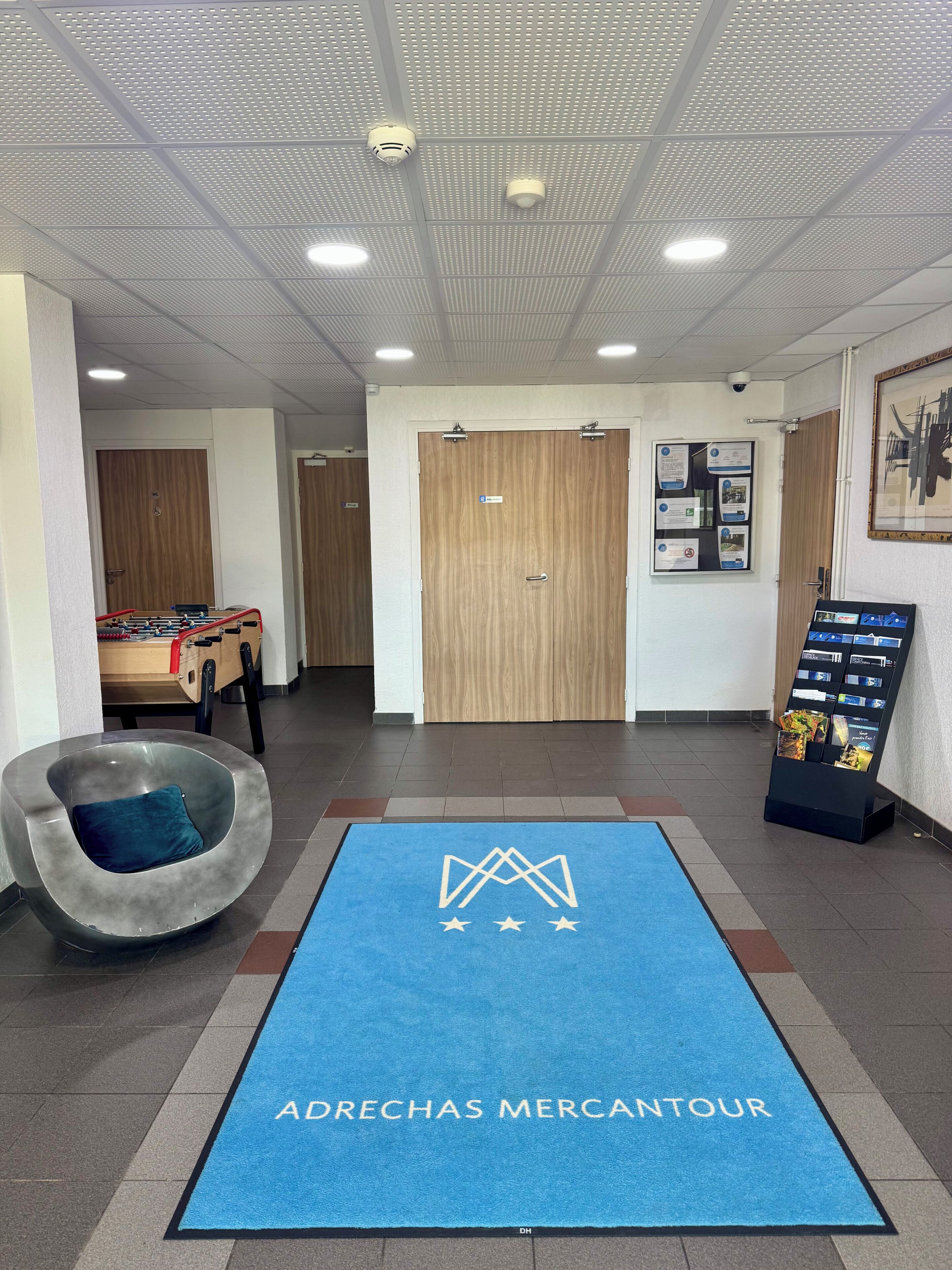 Um saguão de hotel com carpete azul, mesa de futebol de mesa, área de estar diferenciada e um mural de avisos.