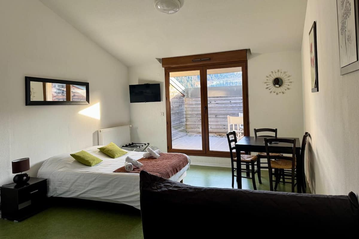 A bedroom with a bed, a desk, chairs, a TV, a mirror, and a view of the outside.