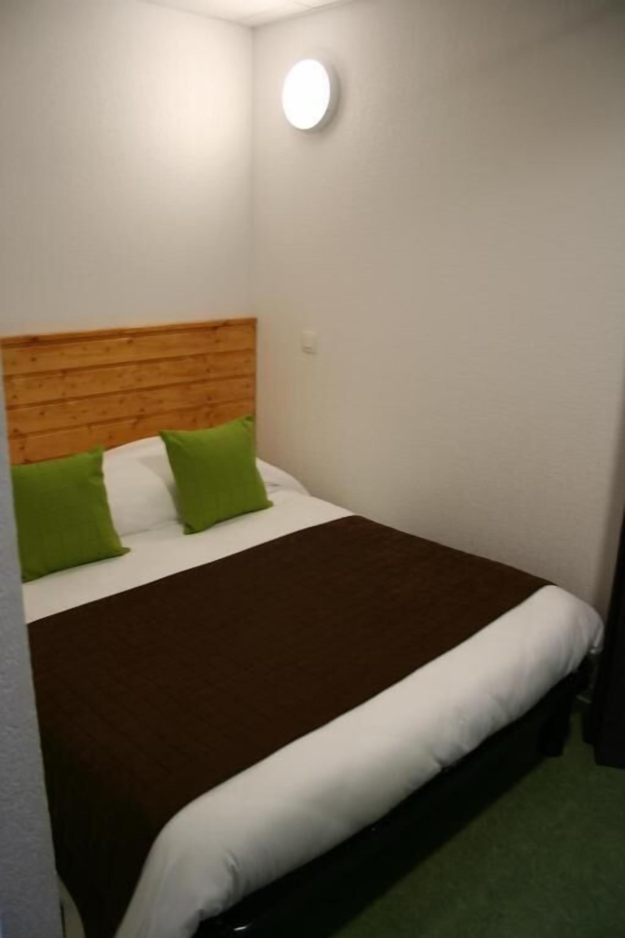 Uma cama de solteiro com cabeceira de madeira, travesseiros verdes e um cobertor marrom em um quarto com paredes brancas e uma luminária redonda.