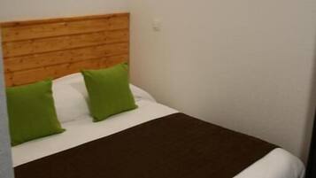 Uma cama de solteiro com cabeceira de madeira, travesseiros verdes e um cobertor marrom em um quarto com paredes brancas e uma luminária redonda.