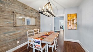 Ein Essbereich mit Holztisch, weißen Stühlen, einer Chandelierleuchtre und einem Gemälde an der Wand.