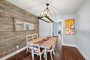 A dining area with a wooden table, white chairs, a chandelier, and a painting on the wall. - Hot Tub Retreat/Walk to Beach/Dual Primary Suites/Monthly Discount (Galveston)