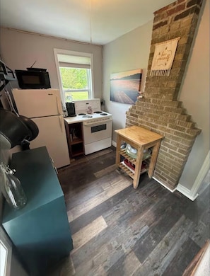 A compact kitchen with a microwave, stove, and refrigerator, a small wooden table, and a brick wall with a painting.