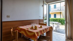 A dining area with a table set for four, a wooden bench, and a view of an outdoor balcony with a white chair and a small table. - Residence Michelangelo two-room apartment B4*+ facing the pool first floor for 4 persons (Bibione)