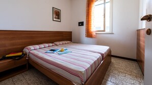 A bedroom with a wooden bed, a bedside table, a window with curtains, and a small framed picture on the wall. - Residence Michelangelo two-room apartment B4*+ facing the pool first floor for 4 persons (Bibione)