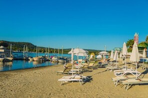 Una playa con tumbonas y sombrillas, barcos amarrados en el agua y un cielo azul despejado.