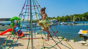Un’area giochi con una struttura a rete per arrampicata, uno scivolo e un’area sabbiosa vicino a un corso d’acqua con barche.
