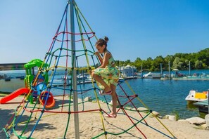 Un’area giochi con una struttura a rete per arrampicata, uno scivolo e un’area sabbiosa vicino a un corso d’acqua con barche.